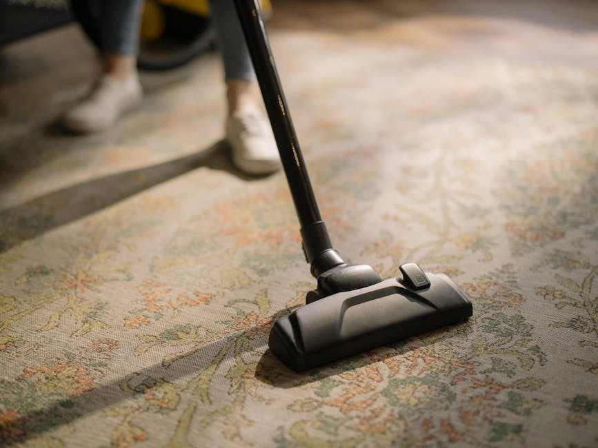 A woman with curly hair, dressed casually in a green jacket, dark jeans, and white sneakers, is performing surface cleaning using a yellow canister vacuum cleaner with a black hose and attachment. She